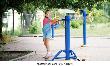 Smiling, Happy Eight Year Old Girl Engaged, Doing Exercises On Outdoor Exercise Equipment, Outdoors, In The Park, Summer, Hot Day During The Holidays. High Quality Photo