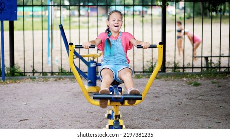 Smiling, Happy Eight Year Old Girl Engaged, Doing Exercises On Outdoor Exercise Equipment, Outdoors, In The Park, Summer, Hot Day During The Holidays. High Quality Photo