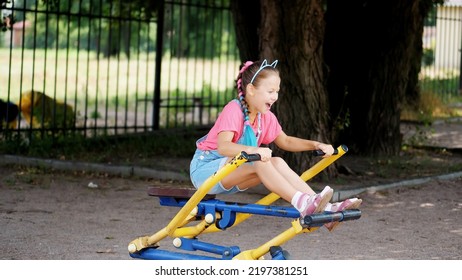 Smiling, Happy Eight Year Old Girl Engaged, Doing Exercises On Outdoor Exercise Equipment, Outdoors, In The Park, Summer, Hot Day During The Holidays. High Quality Photo