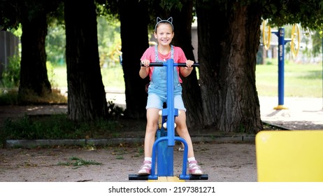 Smiling, Happy Eight Year Old Girl Engaged, Doing Exercises On Outdoor Exercise Equipment, Outdoors, In The Park, Summer, Hot Day During The Holidays. High Quality Photo