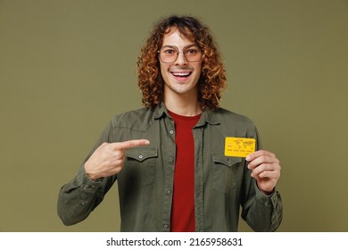 Smiling Happy Fascinating Young Brunet Curly Man 20s Wears Khaki Shirt Jacket Glasses Hold In Hand Pointing Index Finger On Credit Bank Card Isolated On Plain Olive Green Background Studio Portrait