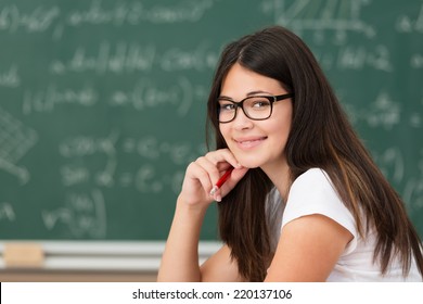 Smiling happy female college student wearing glasses sitting at her desk in the classroom in front of the blackboard smiling at the camera