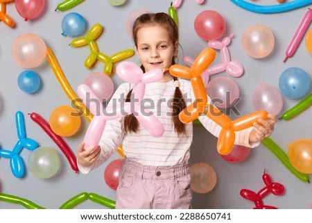 Smiling happy female kid playing at birthday party, cute little girl with braids standing against gray wall decorated with colorful balloons.