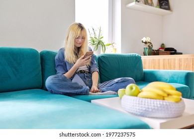 Smiling happy female teenager sitting at home on couch looking at smartphone screen - Shutterstock ID 1977237575