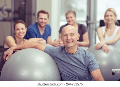 Smiling happy fit senior man in a gym class with a group of diverse people leaning on a pilates ball looking at the camera in a healthy lifestyle concept - Powered by Shutterstock
