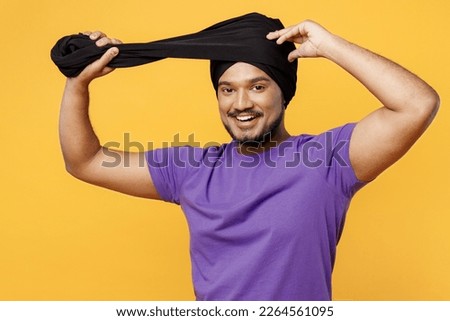 Smiling happy fun devotee Sikh Indian man show how to tie his traditional turban dastar wear purple t-shirt looking camera isolated on plain yellow background studio portrait. People lifestyle concept