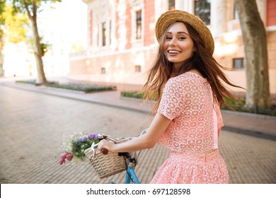 Smiling happy girl in dress and hat riding retro bicycle on a city street and looking at camera