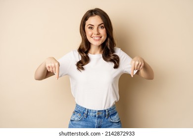 Smiling happy girl pointing fingers down and showing banner, company logo below, click link, standing over beige background - Shutterstock ID 2142872933