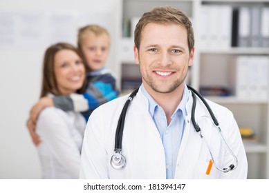 Smiling happy handsome family doctor or GP standing in his surgery with a young mother and child visible in the background