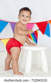 A Smiling, Happy, Healthy, Chubby, Brown Eyed Baby Boy Wearing A Red Cloth Diaper / Nappy Standing On A White Carpet With A Brightly Colored Flag Bunting In The Background.