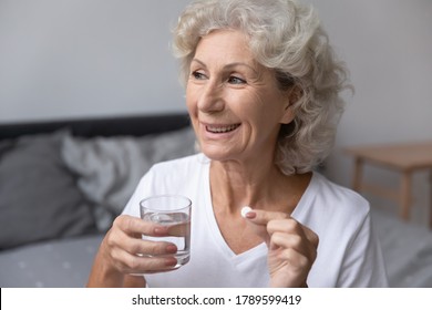 Smiling happy healthy senior woman wake up sits on bed in bedroom alone holding glass of natural water taking daily pill for good health and senile disease prevention, memory meds, vitamins concept - Powered by Shutterstock