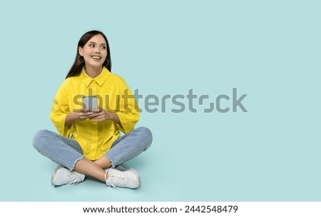 Smiling happy latin young woman in casual outfit sitting on floor, using cell phone isolated on blue studio background, looking at copy space, browsing internet, enjoying newest mobile app