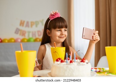 Smiling Happy Little Girl Posing With Cake And Cell Phone On Background, Festive Decorations On Background, Having Video Call, Looking At Device Screen With Smile.