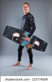 Smiling Happy Man With Short Hair Wearing Wetsuit. Holding Kiteboard. Kite Surfer. Studio Shot. Isolated On Grey Background.