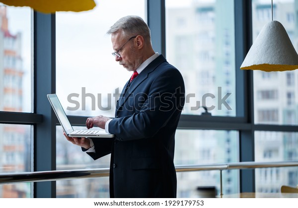 Smiling happy\
managing director thinks about his successful career development\
while standing with a laptop in his office near the background of a\
window with copy space