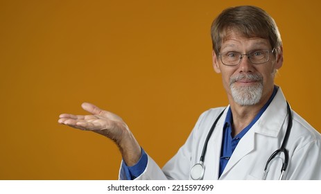 Smiling Happy Mature Bearded Doctor Promoter Hand Out To Copy Space Isolated On Solid Yellow Background Studio. People Sincere Emotions Lifestyle Concept.