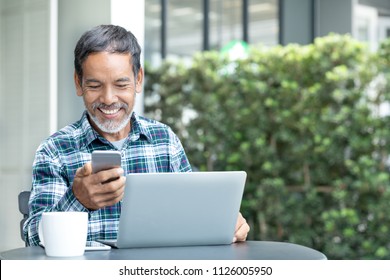 Smiling Happy Mature Man With White Stylish Short Beard Using Smartphone Gadget Serving Internet At Coffee Shop Cafe Outdoor. Laughing Old Man Using Social Media Network Technology Feeling Excited.