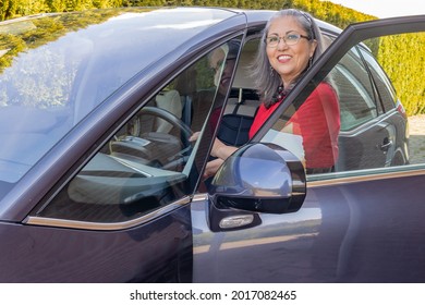 Smiling And Happy Mature Mexican Woman Getting Into Her Gray Car With A Red Dress, Tanned Skin, Gray Hair, Glasses, Black Accessories, Headband With Light Makeup, Sunny Day To Enjoy