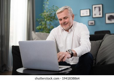 Smiling Happy Middle Aged 50s Old Senior Man Using Laptop Computer Technology Working Online, Surfing Internet, Typing Email, Shopping In Ecommerce Store Relaxing With Device Sitting On Couch At Home.