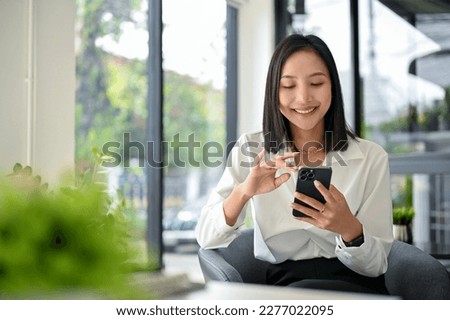 Smiling and happy millennial Asian businesswoman chatting with her friends on her phone, scrolling on social media or watching video on the internet, using her phone while relaxing in the office.
