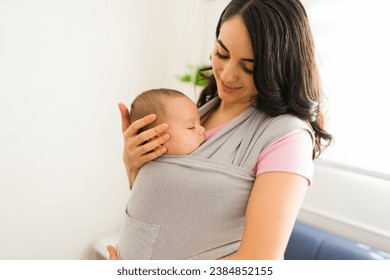 Smiling happy mother feeling a lot of love for her infant child while using a baby sling carrier around the house