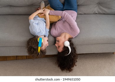 Smiling Happy Mother And Kid Son Lying Upside Down On Couch At Home, Laughing, Tickle Each Other, Hearing Songs In Earphones. Excited Child And Mom Wear Headphones Enjoy Listening To Music Together.