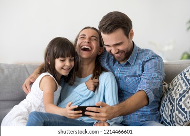 Smiling happy multi-ethnic family sitting on couch in living room at home. Mother father and little daughter laughing using mobile phone watching funny videos online have fun looking humorous program