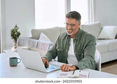 Smiling happy older mature middle aged professional man wearing eyeglasses looking at computer sitting at living room table, using laptop hybrid working, elearning, browsing online at casual home. - Powered by Shutterstock