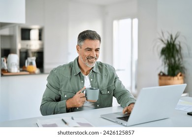 Smiling happy older mature middle aged professional man drinking coffee looking at computer sitting at kitchen table, using laptop hybrid working, elearning, browsing online relaxing at casual home. - Powered by Shutterstock