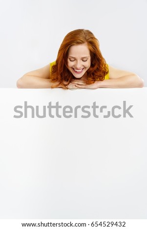 Smiling happy pretty young redhead woman looking down at a blank white sign with copy space as she leans her arms along the top