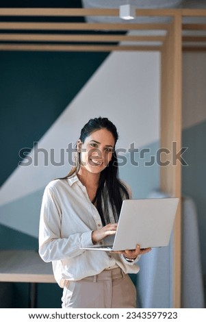 Smiling happy professional young latin business woman company employee, lady executive manager, female worker looking at camera holding laptop standing in modern office, vertical portrait.