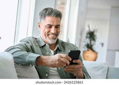 Smiling happy senior mature middle aged man holding cell mobile phone using smartphone sitting at home on couch, scrolling social media, checking financial apps, buying online, texting messages. - Powered by Shutterstock