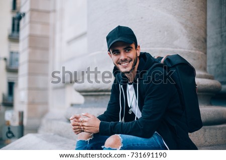 Smiling happy teenager or young male student sits on top of stairs in front of university or highschool, wears black hoodie and cap, smiles at camera, successful and handsome