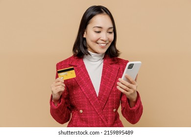 Smiling Happy Woman Of Asian Ethnicity 20s Wear Red Jacket Using Mobile Cell Phone Hold Credit Bank Card Doing Online Shopping Order Delivery Booking Tour Isolated On Plain Pastel Beige Background.