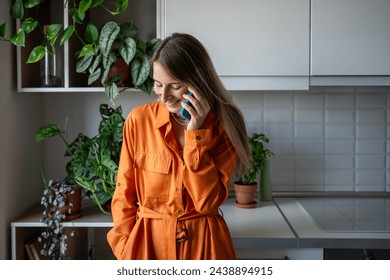 Smiling happy woman enjoy pleasant conversation on cellphone at home. Relaxed Scandinavian middle aged female in orange dress make smartphone call listening funny story. Mobile phone communication.