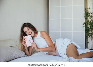 Smiling happy woman with gift box lying in bed and looking at camera - Shutterstock ID 2395492743