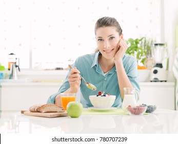 Smiling happy woman having a relaxing healthy breakfast at home sitting at kitchen table, she is eating cereals with fruit and yogurt - Powered by Shutterstock