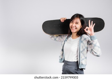 Smiling happy woman holding skateboard on shoulder and show OK sign, Happy Asian beautiful young female excited hold hands longboard, studio shot isolated on white background with copy space