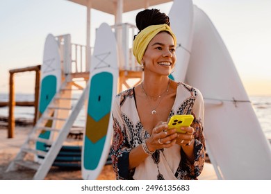 smiling happy woman posing on beach using smart phone dressed in stylish summer outfit colorful yellow boho trend with surfboards on background, people of color - Powered by Shutterstock