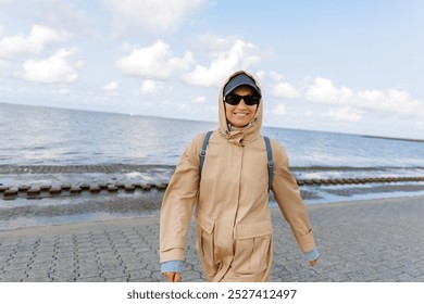 Smiling happy woman walking by Nord sea coast wear beige coat  sunglasses enjoys windy day cloudy sky background. Female person have fun hiking adventure travel at Baltic sea seaside - Powered by Shutterstock