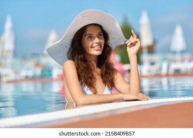 Smiling happy woman wearing trendy white hat standing in swimming pool. Portrait view of beautiful brunette female relaxing in clear pool water in afternoon outdoors. Concept of relaxation. - Powered by Shutterstock