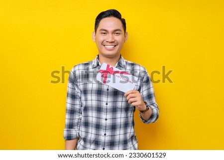 Smiling happy young Asian man wearing a white checkered shirt holding a gift voucher isolated on yellow background. People Lifestyle Concept
