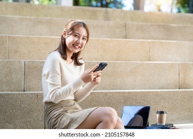 Smiling Happy Young Asian Student Sitting Outside On College Steps Using Mobile Phone. Looking At Camera.