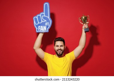 Smiling happy young bearded man 20s in yellow t-shirt cheer up support favorite sport team look camera hold fan foam glove finger up champion cup isolated on plain dark red background studio portrait