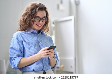 Smiling happy young business woman manager holding smartphone working standing in office using mobile cell phone, checking financial banking apps, browsing online on cellphone typing on cellular. - Shutterstock ID 2220115797