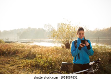 Smiling, happy young Caucasian woman taking a break during early morning mountain bike ride along the lake shore, typing messages on smart phone
