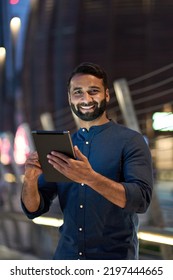 Smiling Happy Young Eastern Indian Business Man Professional Standing Outdoors On Street Holding Using Digital Tablet Online Tech In Night City With Urban Lights Looking At Camera, Vertical Portrait.