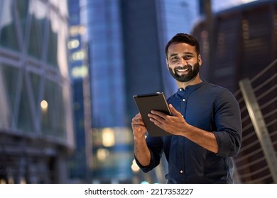 Smiling happy young eastern indian business man professional manager standing outdoor on street holding using digital tablet online fintech in night city with urban lights looking at camera, portrait. - Shutterstock ID 2217353227