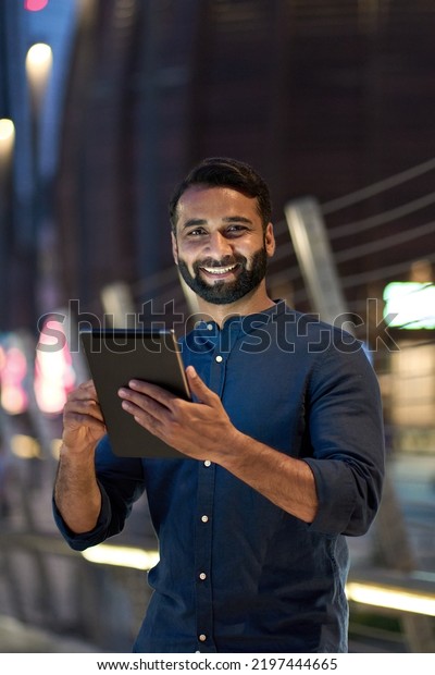 Smiling happy young eastern indian business man\
professional standing outdoors on street holding using digital\
tablet online tech in night city with urban lights looking at\
camera, vertical\
portrait.