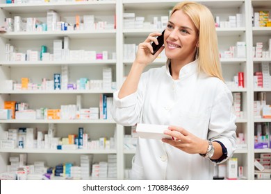 Smiling happy young female pharmacist talking on the phone and holding a box of medications while standing in front of the shelves in pharmacy - Powered by Shutterstock